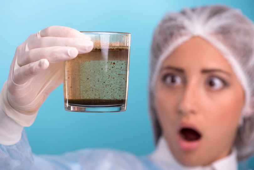 Close-up portrait of woman testing the quality of water in laboratory, holding a glass with bad results and surprised looking on very dirty water with selective focus on a glass, isolated on blue background