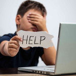 Sad and scared young boy with computer laptop suffering cyberbullying and harassment being online abused by stalker or gossip feeling desperate and humiliated in cyber bullying concept.